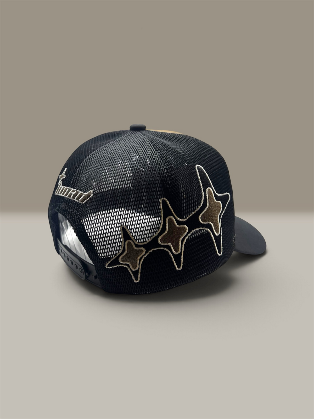 A dark black trucker-style cap with a mesh back, featuring three embroidered star patches outlined in white on the front and a white embroidered script logo on the side, set against a neutral beige background.