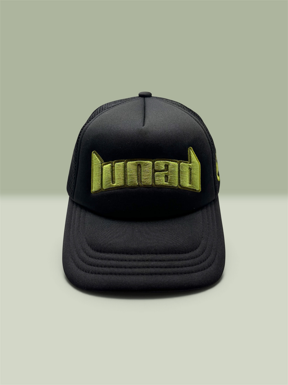 A black trucker-style baseball cap with a curved brim, featuring the word 'Lunad' embroidered in a metallic olive green stylized font across the front. The cap is photographed against a solid, muted sage green background.