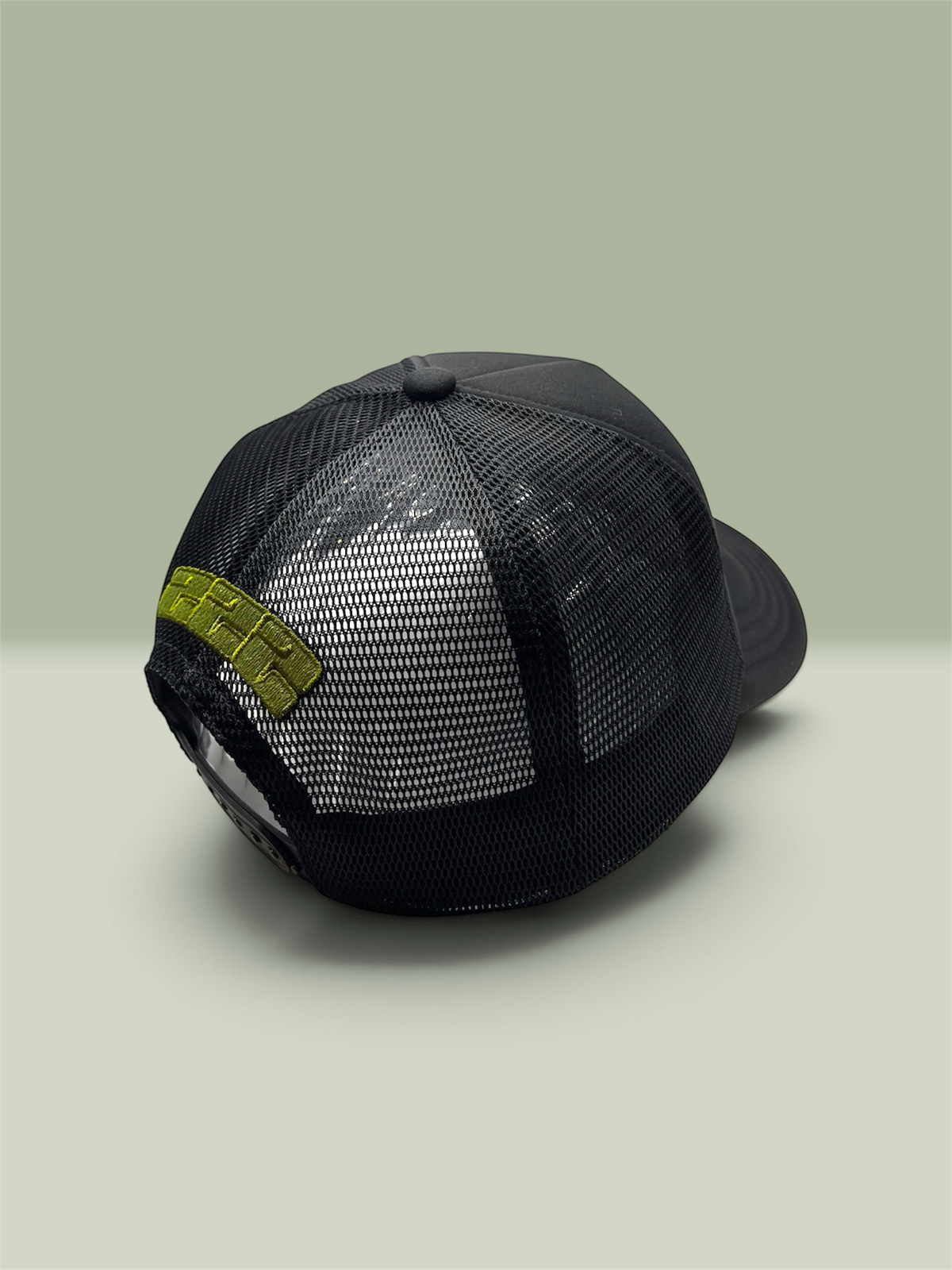 A black trucker-style baseball cap with a mesh back, shown from a three-quarters rear angle. It features a bright sage green rectangular 222 on the rear with the brand name LUNAD 