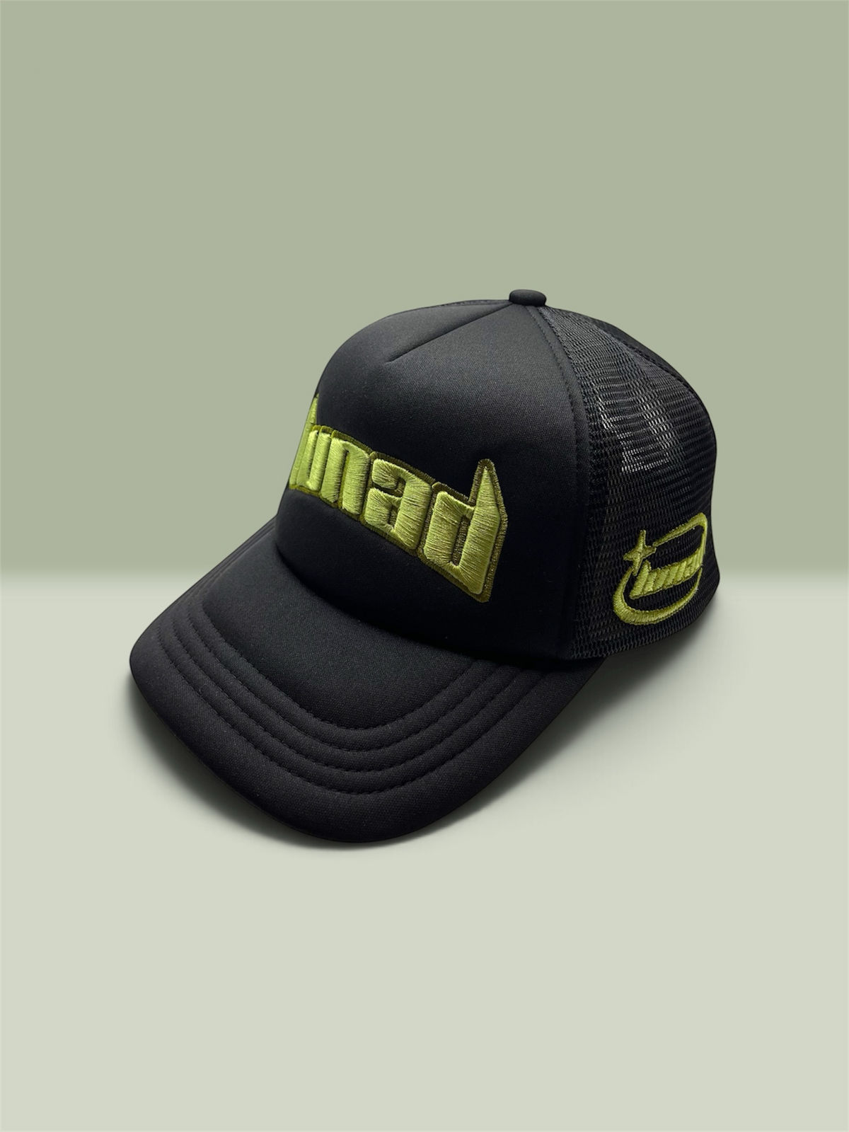 A black trucker-style cap with a curved brim, black mesh back, and bright sage-green embroidered 'Lunad' logos on the front and side, photographed on a light green studio background.