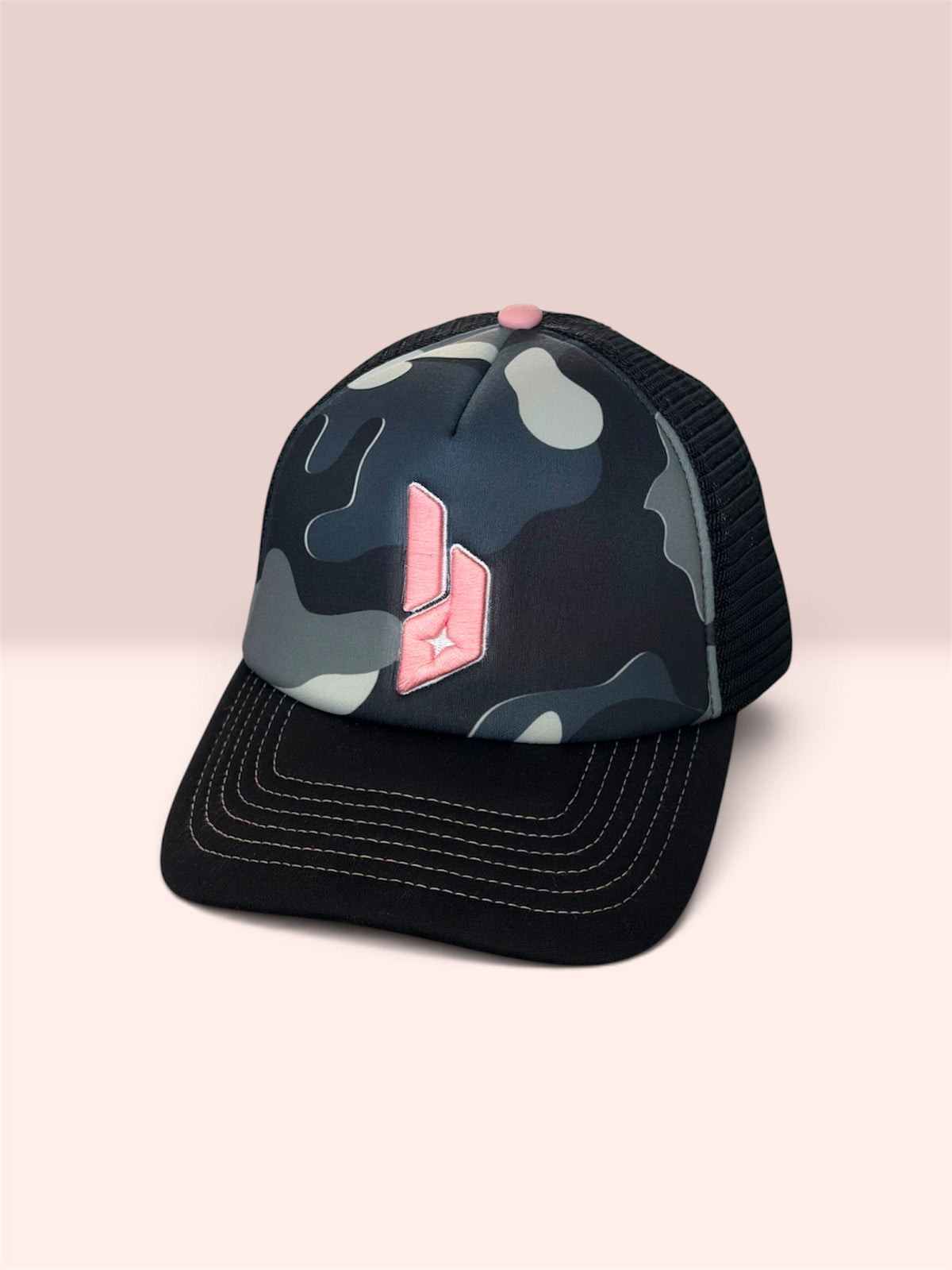 A black trucker cap with a dark grey camouflage front panel and a pink embroidered logo patch with pink stitching, set against a solid pale pink background.