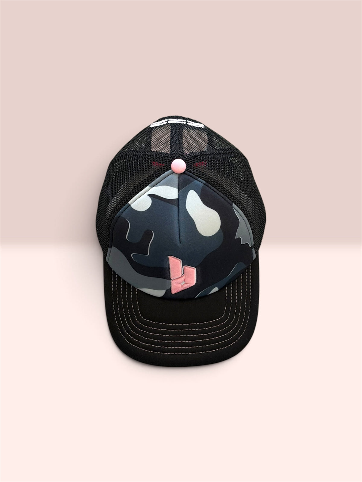 A black trucker cap with a black mesh back, a curved brim, and a front panel featuring a dark camouflage pattern with white patches and a pink graphic accent. Pink stitching. The cap is photographed against a solid pale pink background.