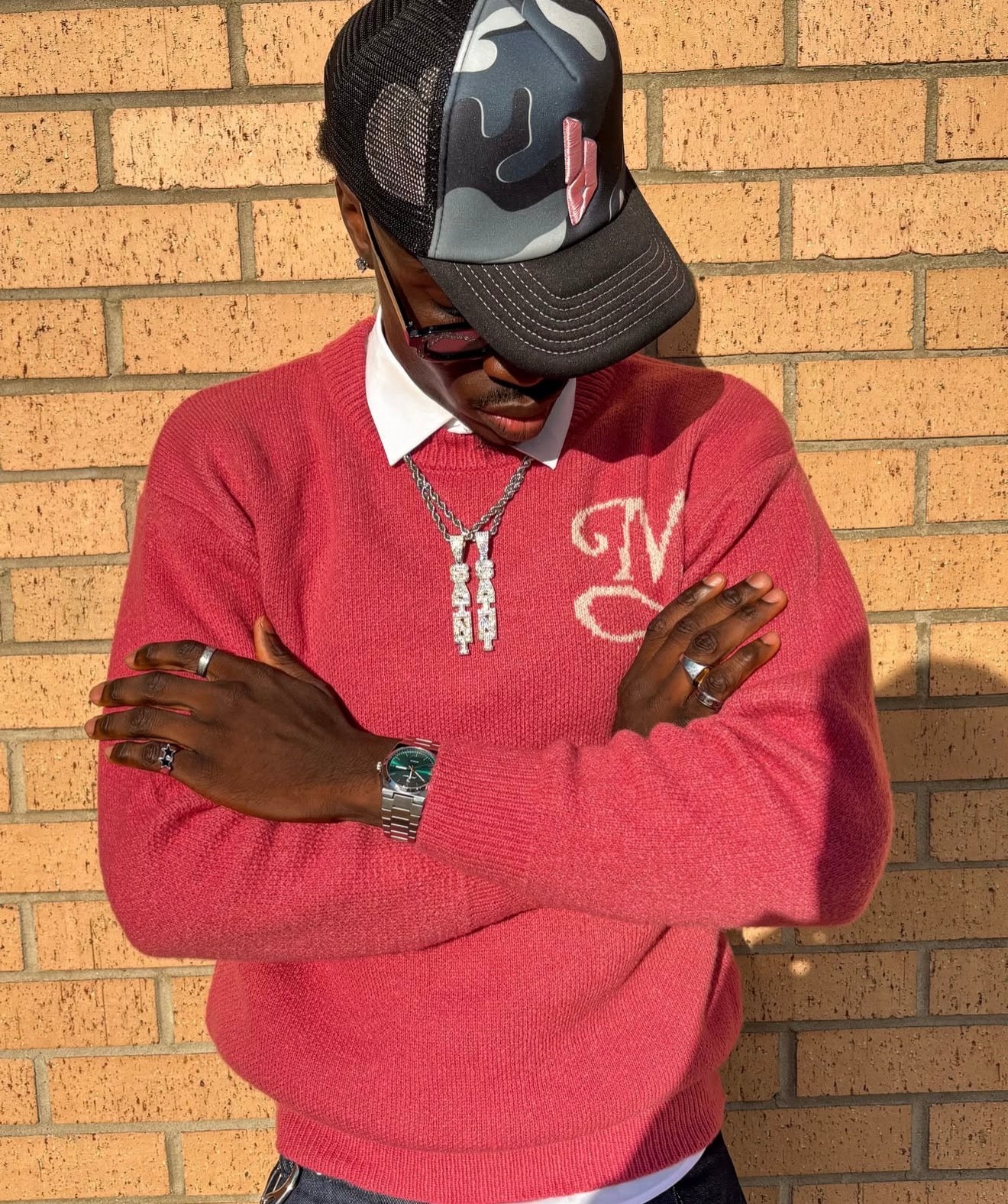 A man wearing a bright pink knit sweater with a white embroidered monogram, a white collared shirt underneath, a silver chain necklace, and a LUNAD black and grey camouflage trucker hat with pink embroidered logo, posing against a tan brick wall.