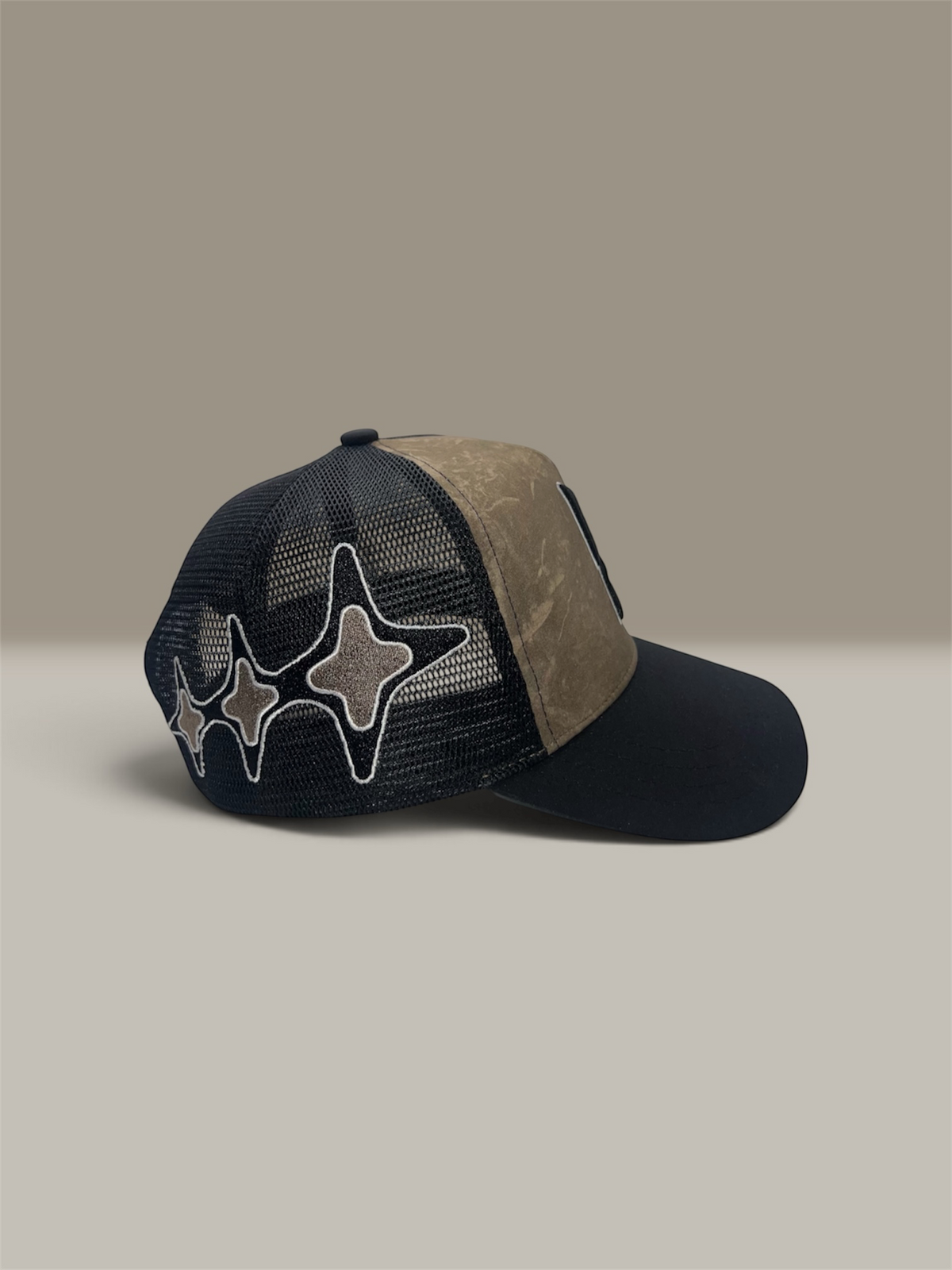 A trucker-style baseball cap with a dark black mesh back, a tree camo tan canvas front panel, and a curved black brim. The front features three large embroidered star-like patches outlined in white.