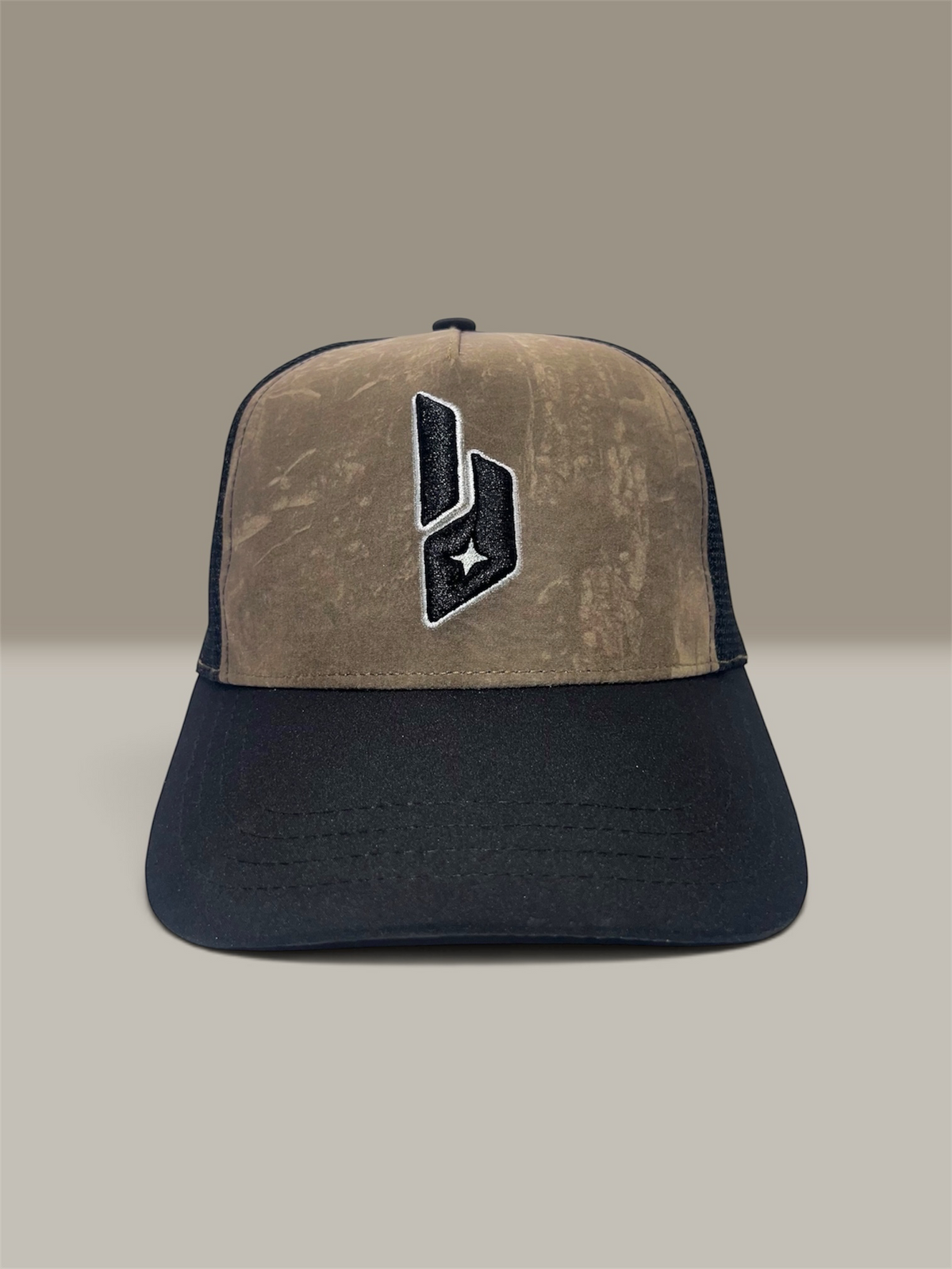 A trucker-style baseball cap with a tree camo tan front panel and a dark black curved brim, featuring a black and white embroidered logo on the front. The mesh back is dark black.